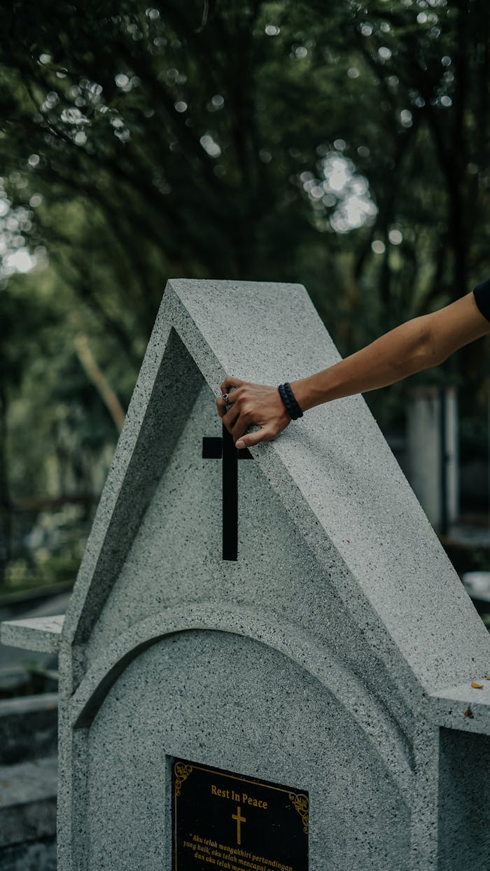 A hand touches a granite tombstone in a tranquil cemetery, framed by lush greenery.