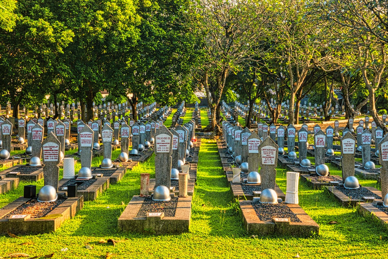 Rows of tombstones with military helmets located on grassy main heroes cemetery with tall lush trees and bright sunlight in Kalibata